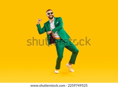 Happy carefree handsome young man in trendy green suit and sunglasses celebrating St Patrick's Day, having fun and dancing on yellow colour studio background. St Patrick's Day and fashion concept