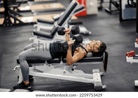 A strong muscular sportswoman is lying on a bench in gym and lifting dumbbells while doing exercises for biceps. Royalty-Free Stock Photo #2259753429