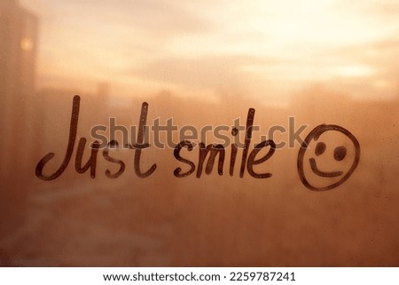 Handwritten lettering text Just smile and happy smile painted on sunset window flooded with raindrops on orange sunset glass, concept photo