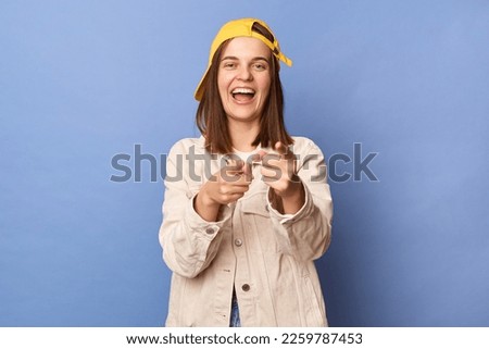 Photo of cute charming positive brown haired teenager girl wearing baseball cap and jacket posing isolated over blue background, pointing at camera, choosing you.