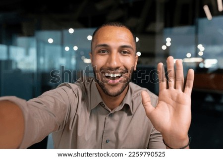 Smiling and cheerful businessman in office taking selfie photo on phone and talking on video call with colleagues and friends using smartphone, african american man waving at camera greeting gesture.