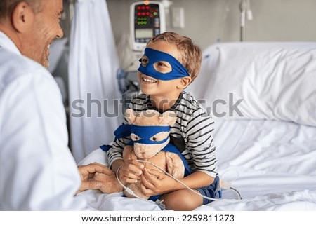 Little boy with superhero mask embracing teddy bear while playing with doctor and sitting at hospital. Playful child dressed as superhero talking to pediatrician. Doctor visiting his little patient. Royalty-Free Stock Photo #2259811273