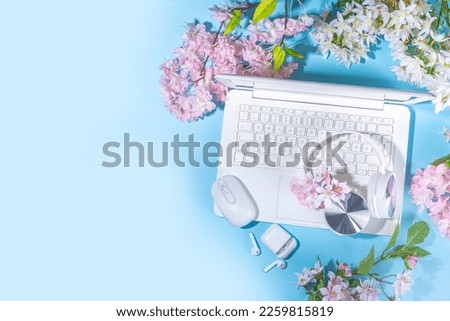Spring office workplace, blogging flat lay background. White laptop, with headphones, tablet, spring flowers bouquet on light blue background, girl's hands typing on a laptop.