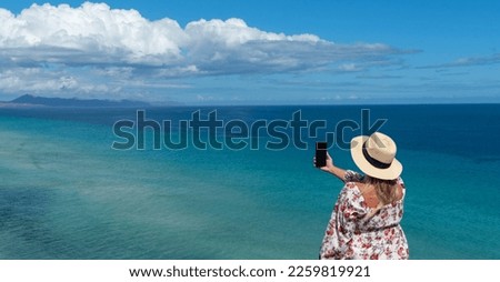 Panoramic view of a blonde woman in a straw hat taking a picture of the turquoise sea off the coast of the tourist island of Fuerteventura in the Canary Islands.