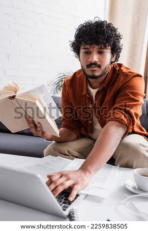 Young indian copywriter holding book and using blurred laptop on couch