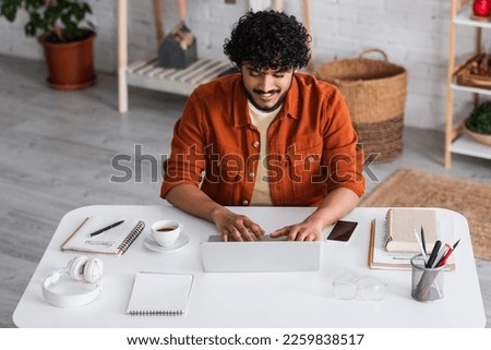 Smiling indian copywriter using laptop near notebooks and coffee at home