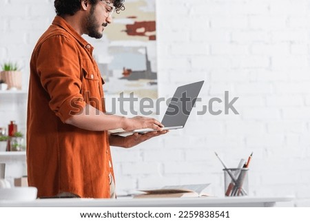 Side view of indian copywriter in eyeglasses holding laptop with blank screen at home