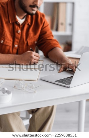Cropped view of indian copywriter using laptop and writing on notebook near eyeglasses at home