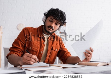 Indian copywriter in headphones holding papers and writing on notebook at home