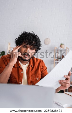 Focused indian freelancer looking at laptop and holding paper a home