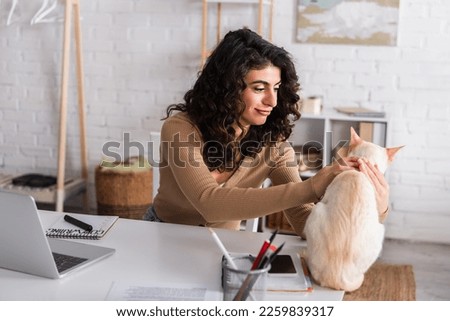 Positive copywriter petting oriental cat near notebooks and laptop at home