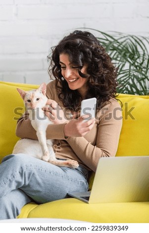 Smiling copywriter holding cellphone and oriental cat near laptop on couch