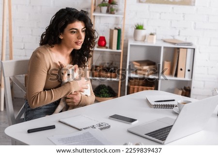 Brunette woman holding oriental cat near gadgets and notebooks at home