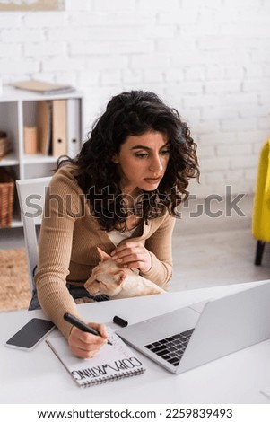 Brunette copywriter with oriental cat holding marker near notebook and devices at home
