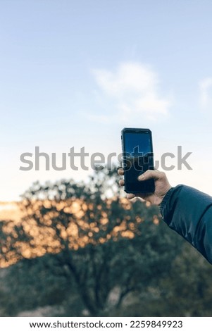 hand of young digital nomad adventurer taking a picture with his smart phone to a mountainous landscape during a clear sunset in vertical
