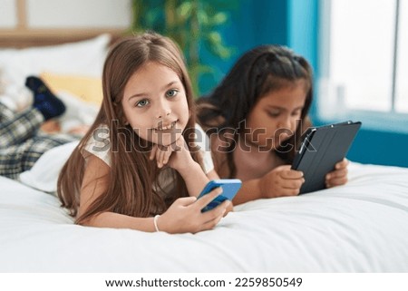 Two kids using smartphone and touchpad lying on bed at bedroom