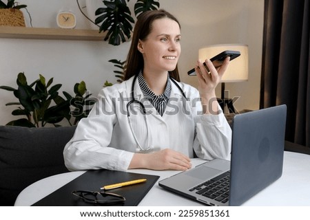 Smiling beautiful young woman doctor recording audio message on smartphone sitting at table with laptop, chatting with patient online, giving consultation or advice, treatment, using virtual voice app Royalty-Free Stock Photo #2259851361