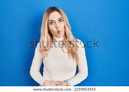 Young caucasian woman standing over blue background making fish face with lips, crazy and comical gesture. funny expression. 