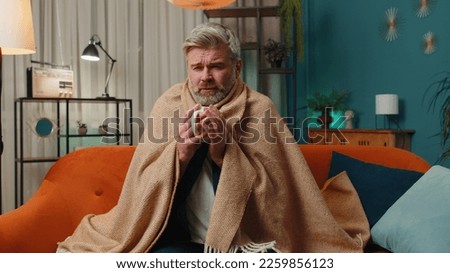 Sick man wrapped in plaid sit alone shivering from cold on sofa drinking hot tea in unheated apartment without heating due debt. Unhealthy senior guy feeling discomfort try to warm up at evening home Royalty-Free Stock Photo #2259856123