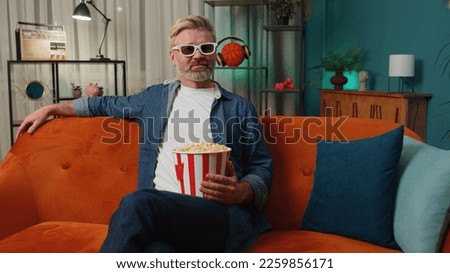 Excited bearded middle-aged man sits on sofa eating popcorn snacks and watching interesting TV serial, sport game, film, online social media movie content at home. Guy enjoying evening entertainment