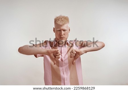 Front view of confident albino european guy showing thumbs down gesture and looking at camera. Young bearded blonde man of zoomer generation wearing t-shirt. White background. Studio shoot. Copy space Royalty-Free Stock Photo #2259868129