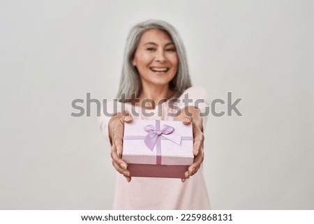 Selective focus of gift present in hands of blurred smiling senior caucasian woman. Elderly female person looking at camera. Holiday and event. Isolated on white background. Studio shoot. Copy space
