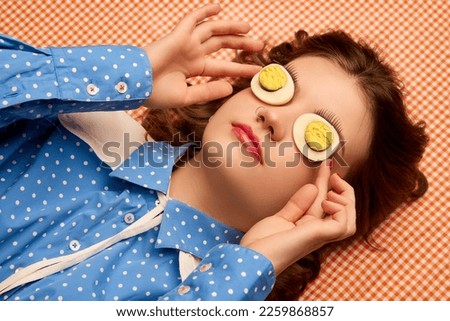 Strange look. Weird people concept. Young girl lying on table with eggs on her eyes over peach color background. Vintage, retro style. Food pop art photography, fashion, kitsch concept Royalty-Free Stock Photo #2259868857