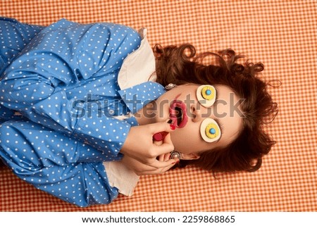 Doing makeup. Weird people concept. Young girl lying on table with eggs on her eyes over peach color background. Vintage, retro style. Food pop art photography, fashion, kitsch concept