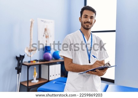 Young hispanic man wearing physiotherapist uniform writing on clipboard at rehab clinic Royalty-Free Stock Photo #2259869203