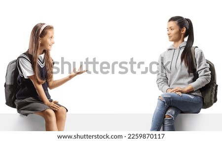 Female pupil talking to an odler female student and sitting on a blank panel isolated on white background