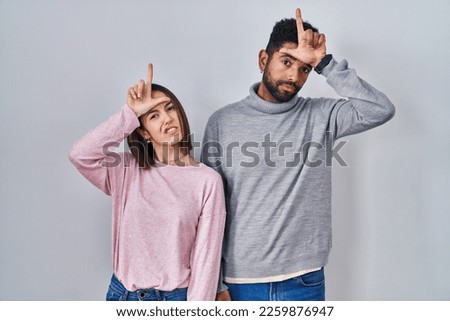 Young hispanic couple standing together making fun of people with fingers on forehead doing loser gesture mocking and insulting. 