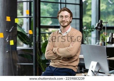 Portrait of successful smiling man inside office, blond man with crossed arms smiling and looking at camera at workplace with computer, businessman happy with achievement result.