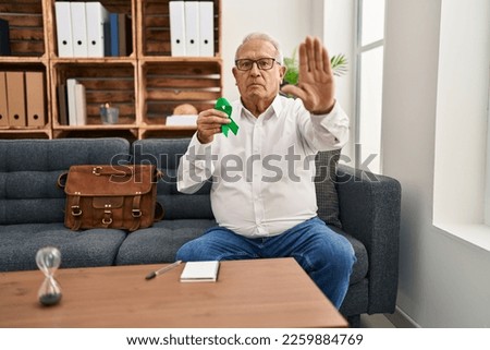 Senior therapist with grey hair holding support green ribbon for mental health awareness with open hand doing stop sign with serious and confident expression, defense gesture 