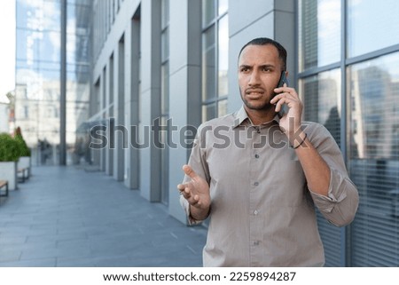 Outdoor portrait of angry and upset man outside office building, businessman in shirt talking on phone and gesturing with hands. Royalty-Free Stock Photo #2259894287