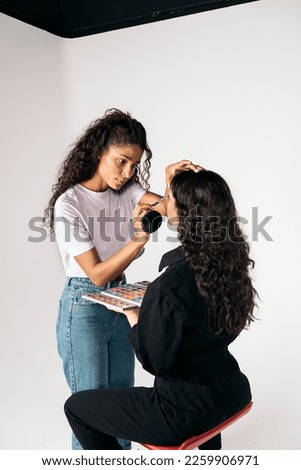 Stock photo of young latin woman doing make up to her client in studio over white background.