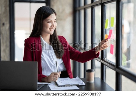 Smart asian business woman mentor coach leader writing idea or task on post it sticky notes on glass wall.