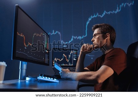 Side view of concentrated crypto trader sitting in front of computers, making professional analysis of candlestick chart, creating strategy, holding pen and notepad, looking at monitor, touching chin Royalty-Free Stock Photo #2259909221