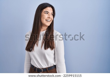 Young brunette woman standing over blue background looking away to side with smile on face, natural expression. laughing confident.  Royalty-Free Stock Photo #2259915115
