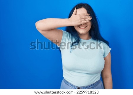 Young modern girl with blue hair standing over blue background smiling and laughing with hand on face covering eyes for surprise. blind concept. 