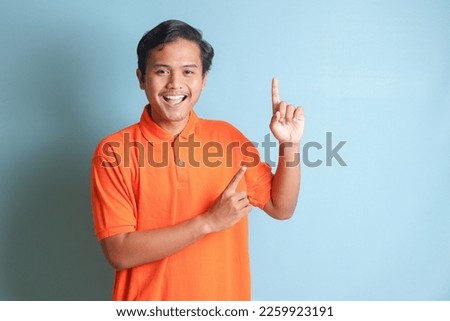 Portrait of excited Asian man in orange shirt smiling and looking at the camera pointing with two hands and fingers to the side. Isolated image on blue background