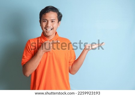 Portrait of excited Asian man in orange shirt smiling and looking at the camera pointing with two hands and fingers to the side. Isolated image on blue background