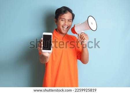 Portrait of attractive Asian man in orange shirt speaking louder using megaphone, promoting product while showing blank screen mobile phone. Advertising concept. Isolated image on blue background