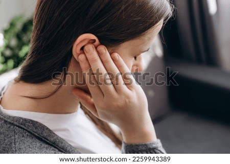 Close up of young brunette female holding painful ear, suddenly feeling strong ache. Unhealthy caucasian woman 20s suffering from painful otitis sitting on couch at home. Health problems concept Royalty-Free Stock Photo #2259943389