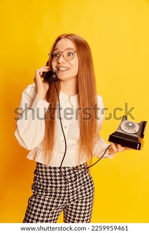 Young happy girl with red hair wearing office style clothes talking on retro vintage phone over yellow background. Concept of youth, student college life, business and education. Model looks happy