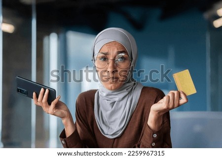 Close up photo. Portrait of a young Arab woman sitting in the office and holding a credit card in her hands. Problems with the account, money. He looks worriedly at the camera, spreads his hands.