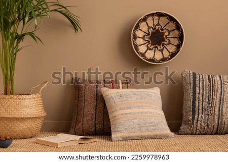 Minimalist composition of ethno living room interior with patterned, colorful pillows, brown wall, braided carpet, round basket on the wall and personal accessories. Home decor. Template.  Royalty-Free Stock Photo #2259978963