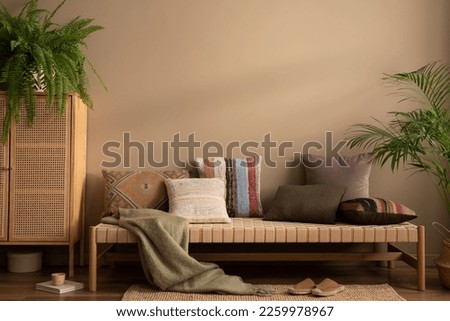 Ethno composition of living room interior with couch, patterned pillows, rattan sideboard, braided rug, fern, plants in flowerpots, brown slippers, cup and personal accessories. Home decor. Template. Royalty-Free Stock Photo #2259978967