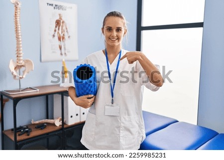 Young physiotherapist woman holding foam roll at the clinic pointing finger to one self smiling happy and proud 