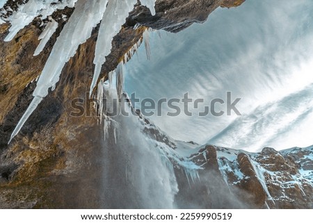 The frozen Kvernufoss waterfall, with snow and bluish stalagmites with water falling from the view like a cave and cobweb cloudy sky in Iceland