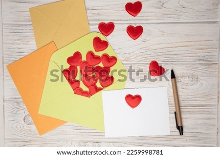Light green, orange, yellow envelopes and a white card for text with red falling hearts on a white wooden background. Happy Valentine's Day greeting, background. Flat lay